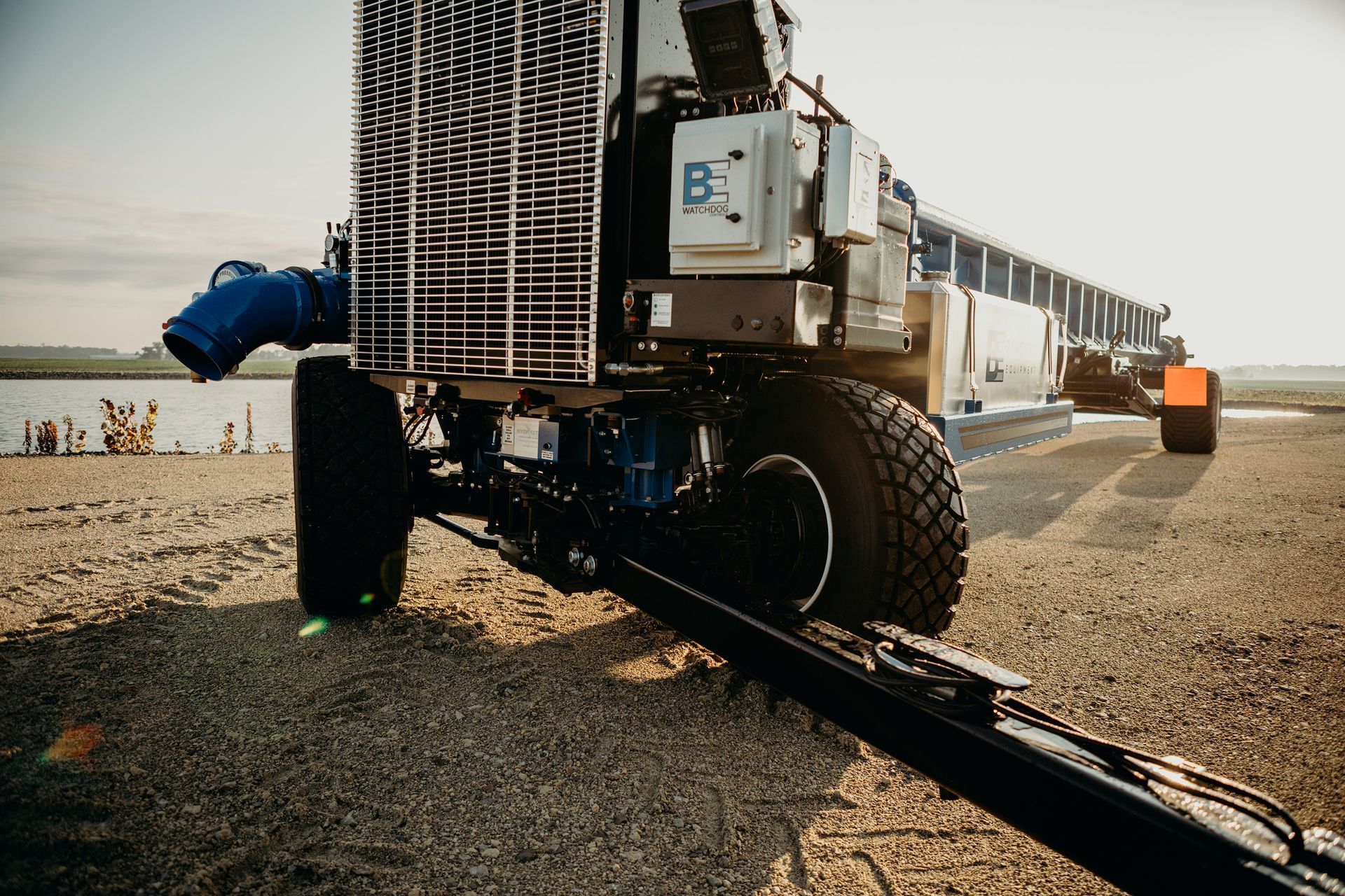 Large industrial machine with oversized tires on a gravel path near the shore, extending equipment to the right.