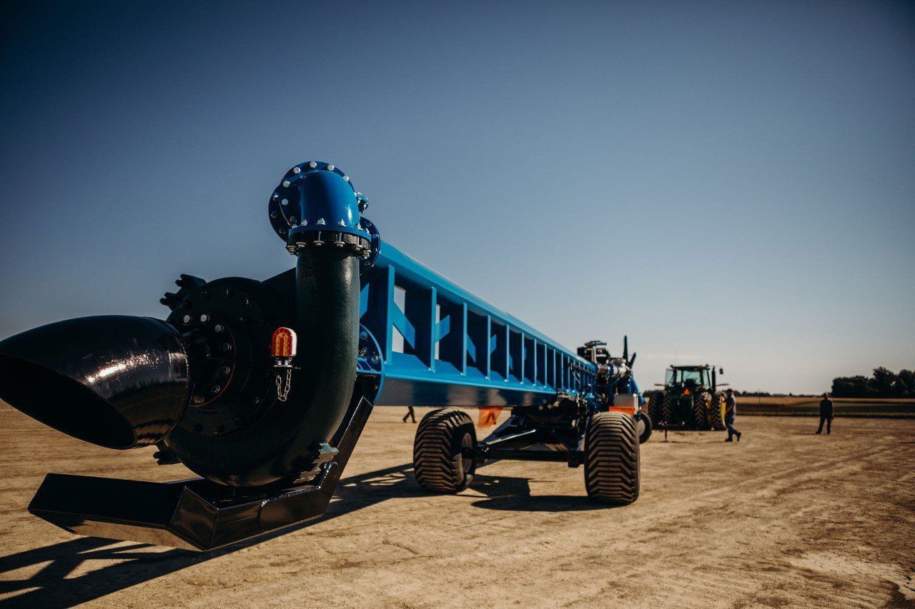 Blue farm implement with large black blade and wheels on a dusty field under a clear sky