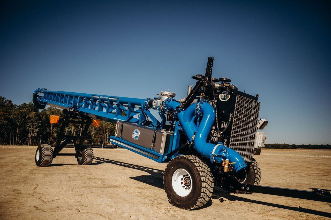 Blue agricultural machine with a long conveyor on a dirt field under a clear sky