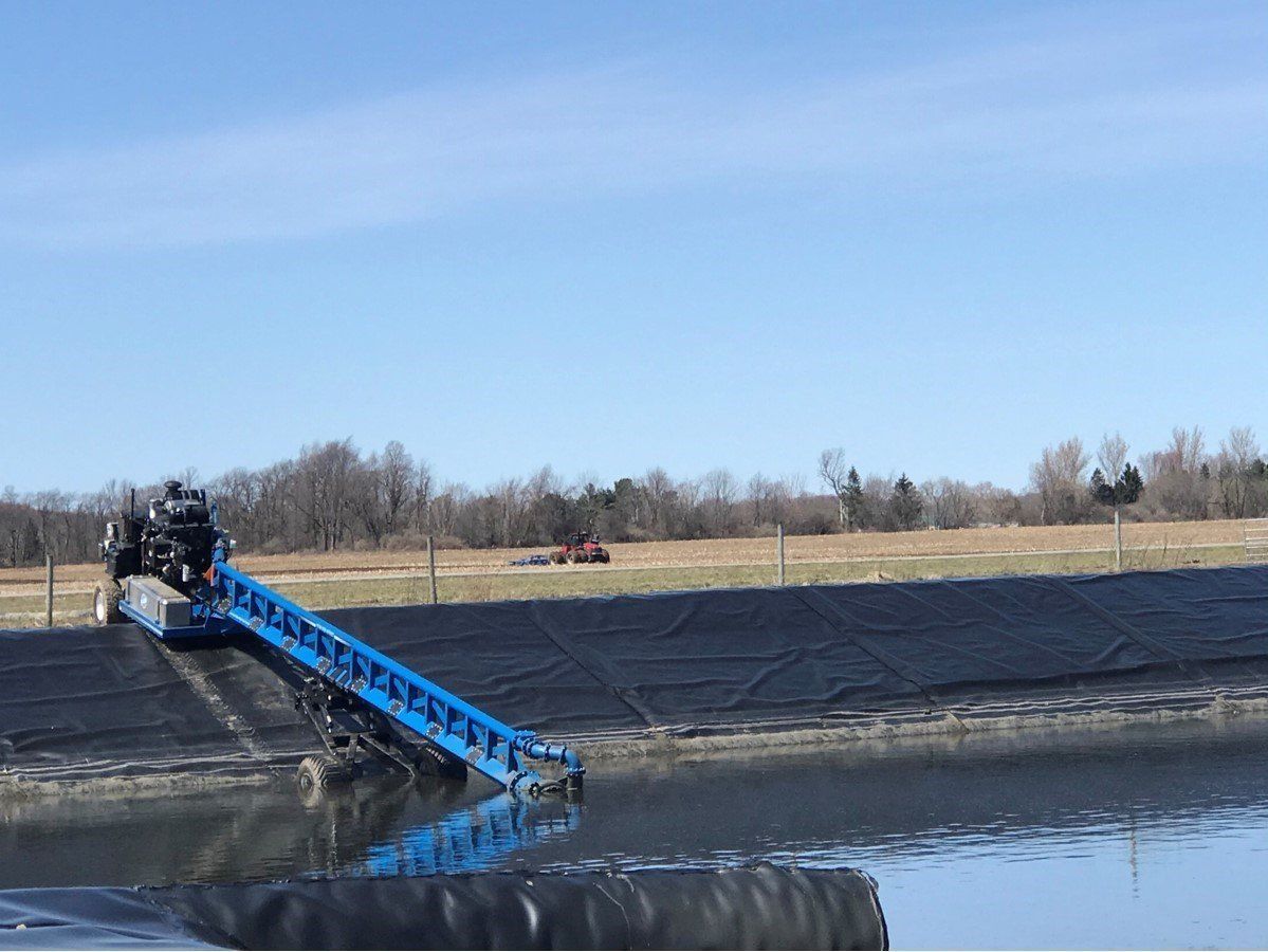 Blue conveyor working over a pond to spread black liner material under a clear sky