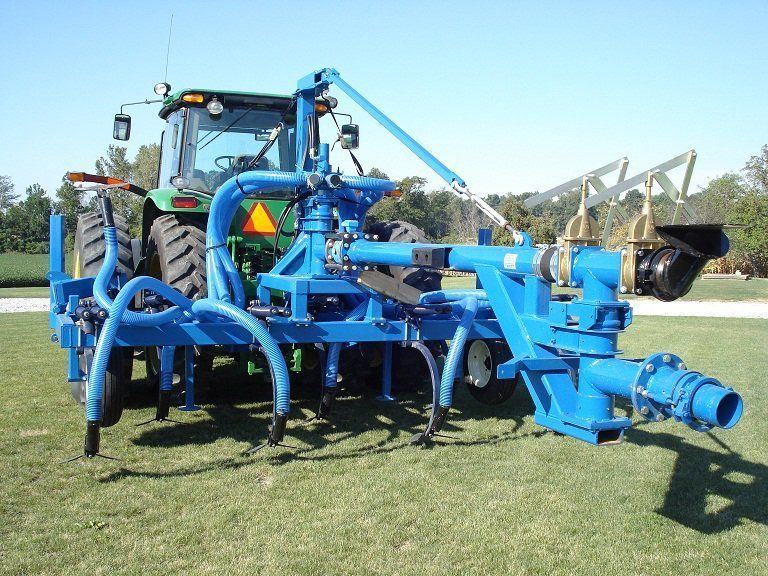 Blue agricultural machine attached to a tractor on a grassy field, with large tines and hoses.
