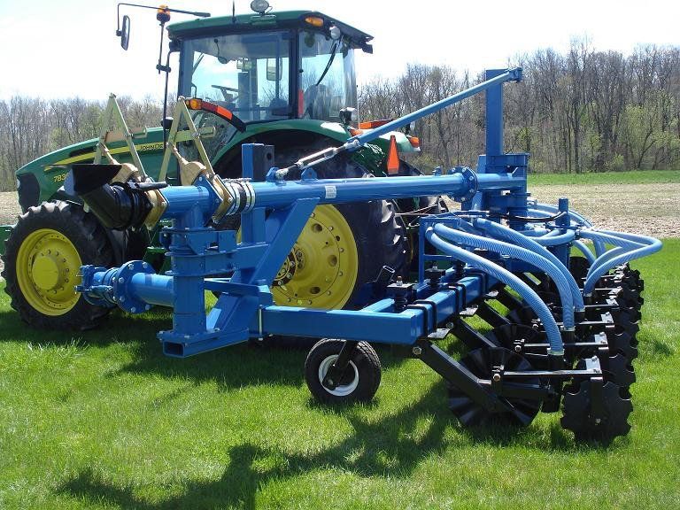 Blue agricultural tillage implement attached to a tractor in a field, with curved tines and yellow wheels