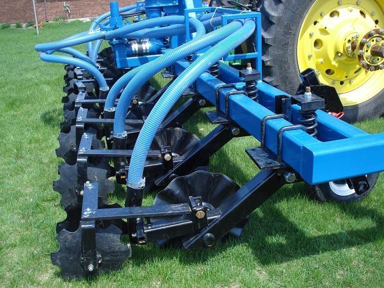 Blue agricultural tillage implement attached to a tractor on grass, with curved hoses and metal tines