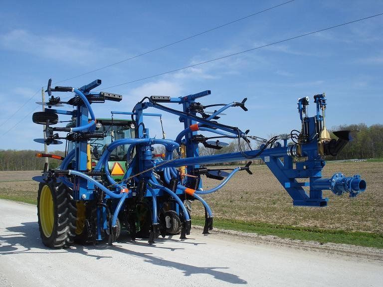 Blue farm sprayer with folded booms on a gravel road beside a field under a clear sky