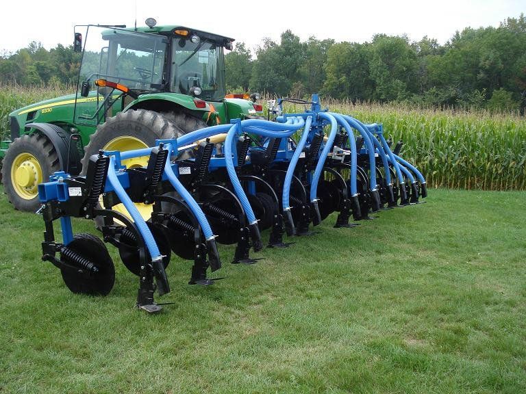 Green tractor pulling a blue crop tillage implement across a grassy field beside corn rows