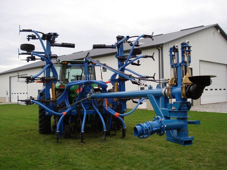 Blue agricultural machine with multiple arms and tines on a grassy field beside a white building