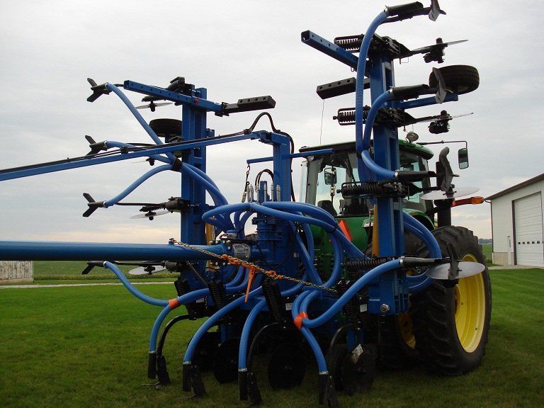 Blue farm spraying machine with multiple booms parked in a field under a cloudy sky