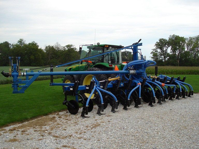 Blue agricultural tillage machine with curved tines parked on a gravel path beside a grassy field