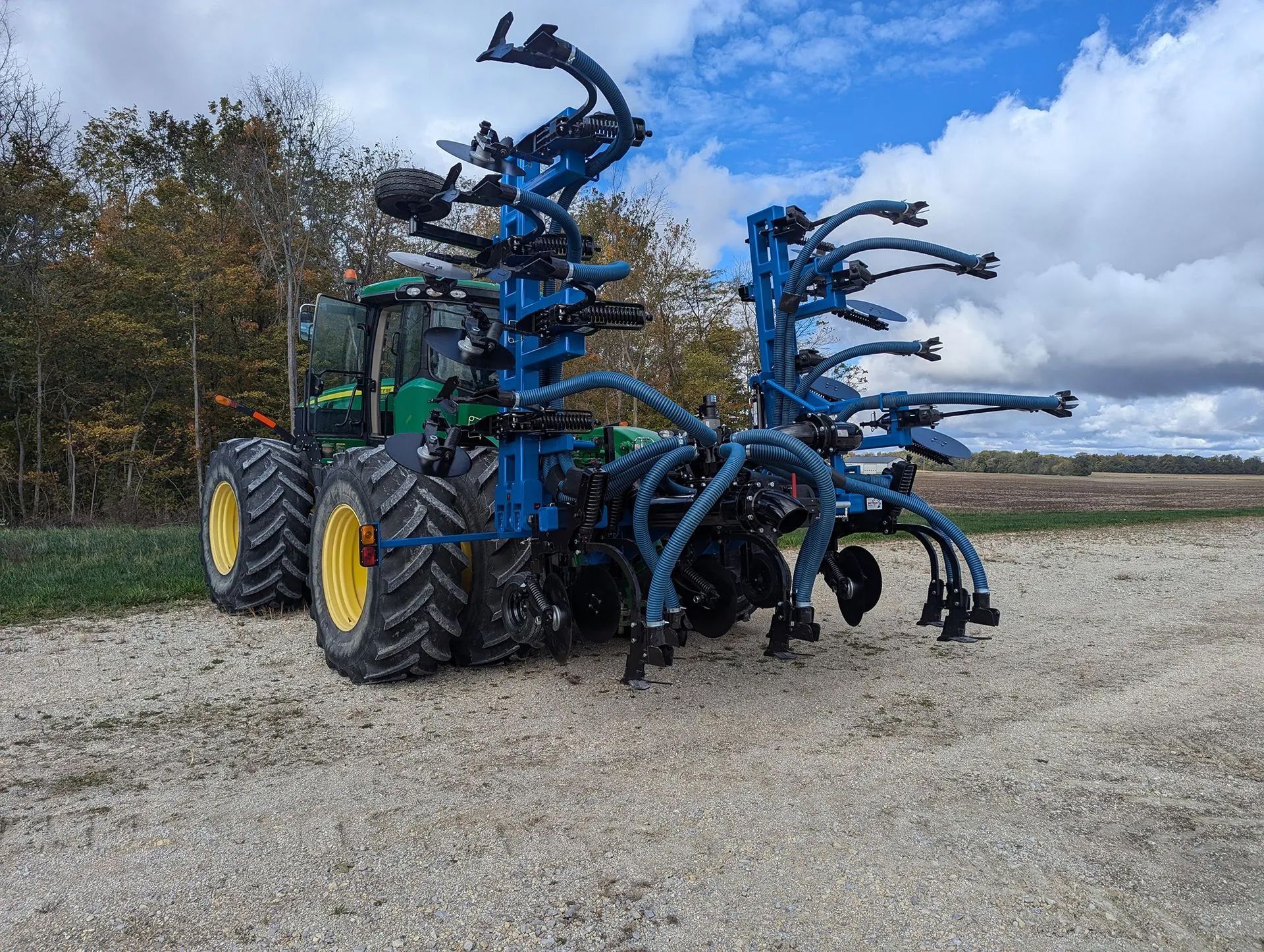 Large blue agricultural machine with black hoses parked on a gravel field under cloudy skies