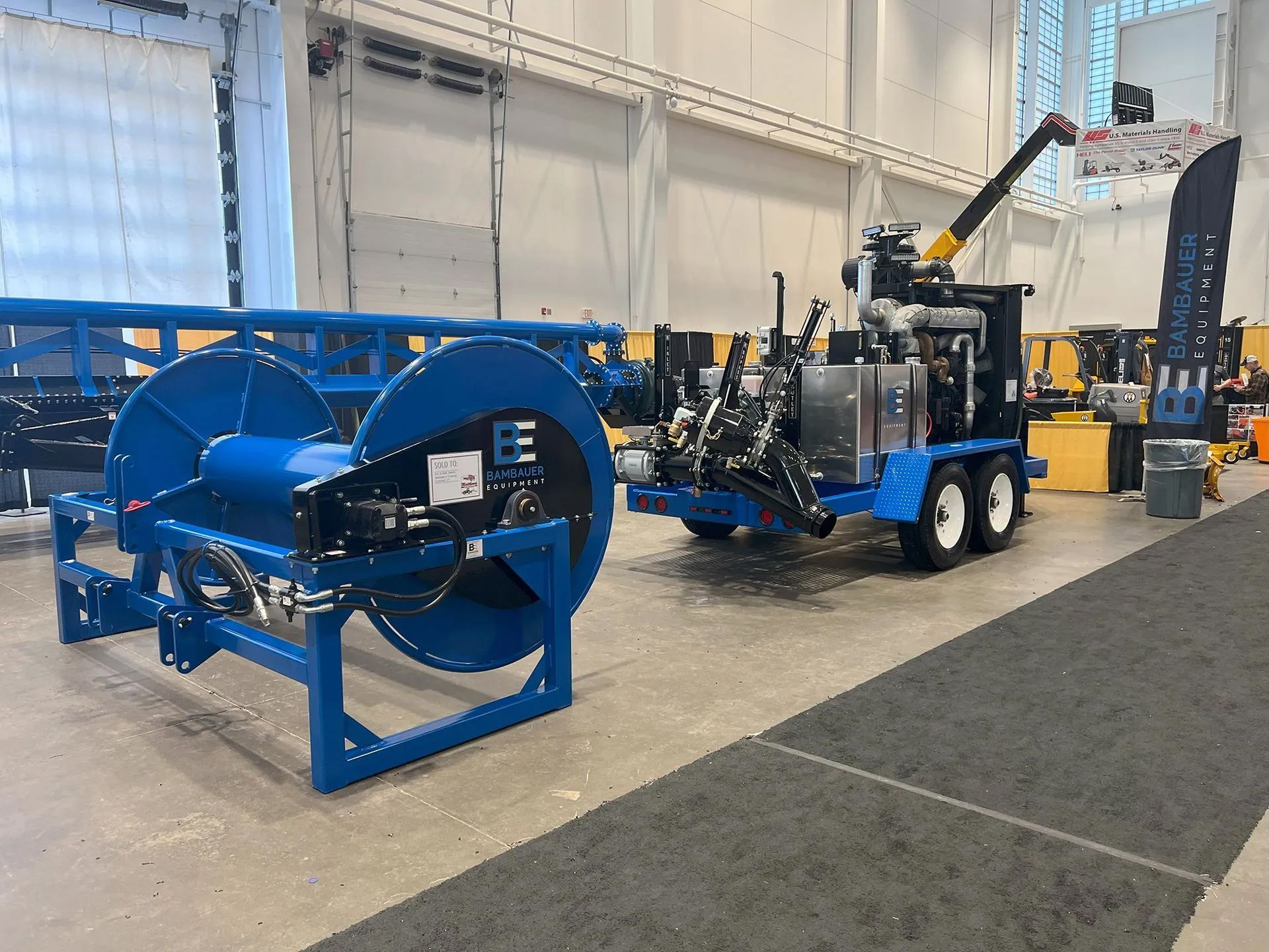 Blue industrial hose reel and trailer-mounted machine displayed indoors at an exhibition hall.