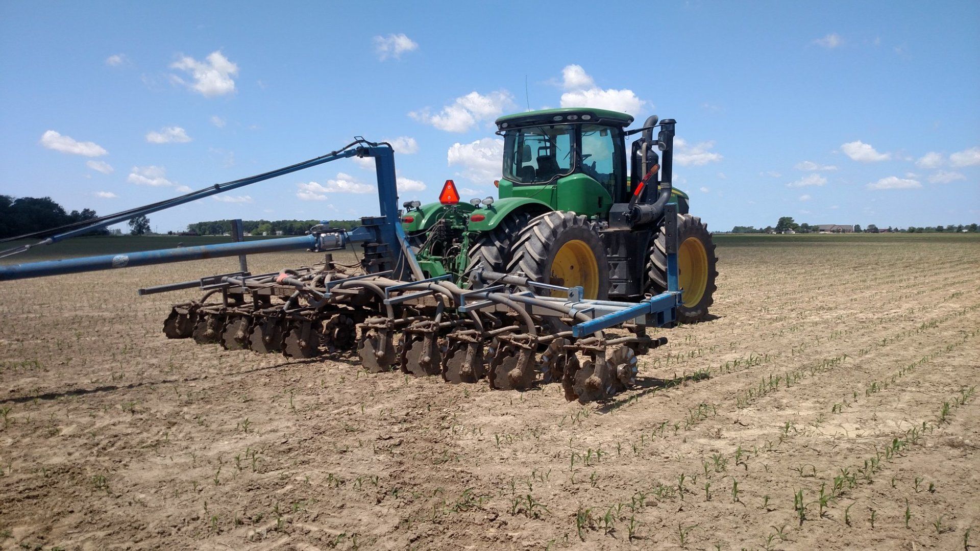 Green tractor towing a wide cultivator across a dry field under a blue sky