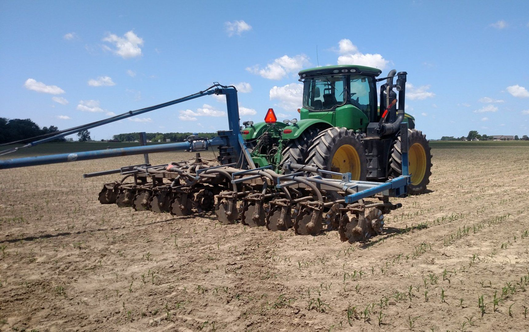 Green tractor pulling farm tillage equipment across a dry field under a blue sky