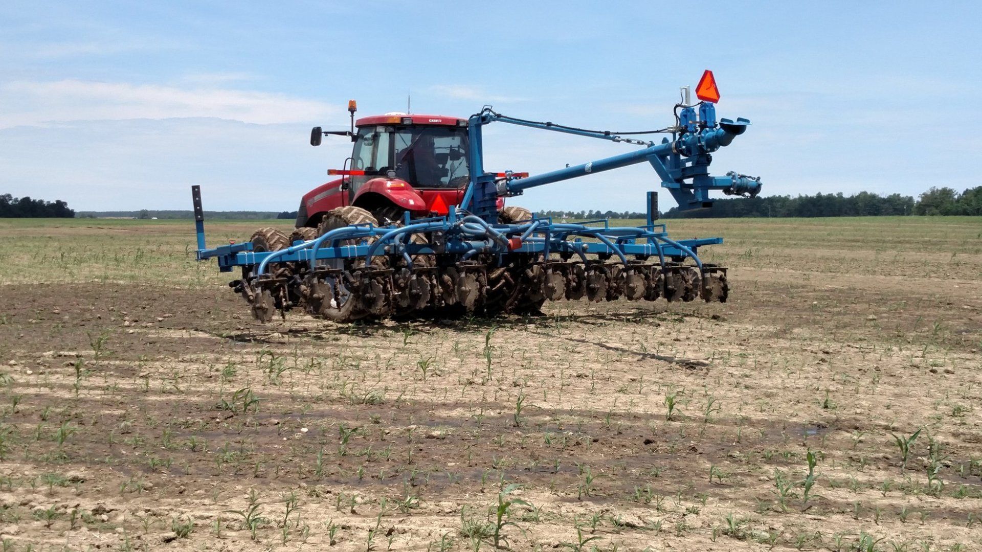 Red tractor pulling a blue farm implement across a dry field under a clear sky