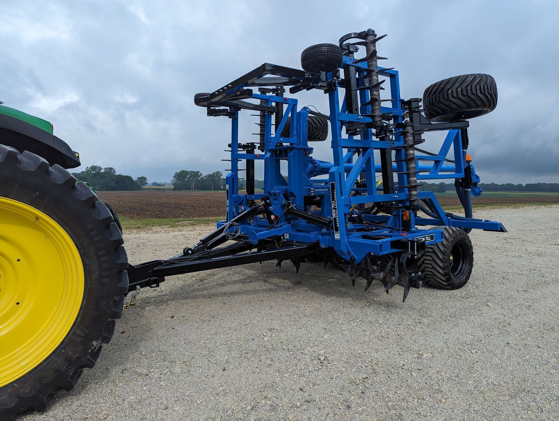 Blue agricultural tillage machine with large black tires and a yellow tractor wheel in a gravel field under cloudy sky
