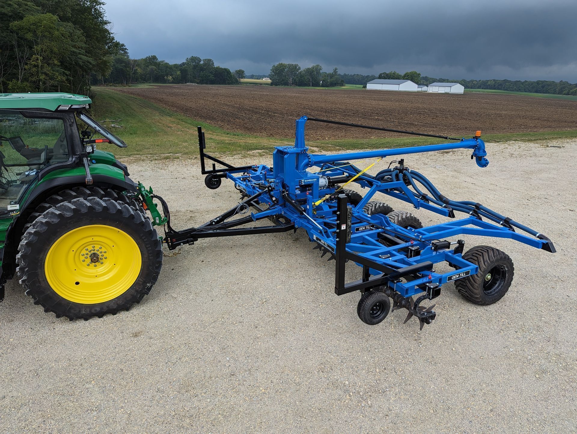 Green tractor towing blue farm implement on a gravel road beside a field