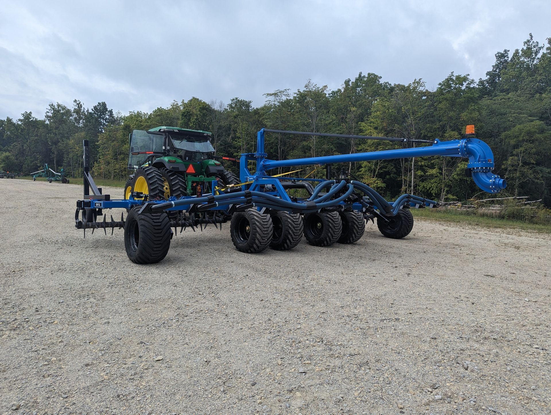 Blue agricultural planter on a gravel road with a tractor attached, in a wooded rural area