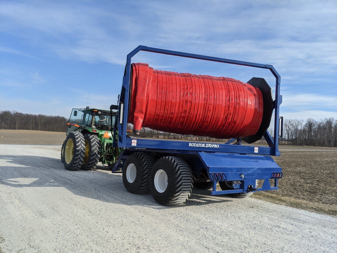 Tractor hauling a large red wrapped hay bale on a blue trailer in a gravel field