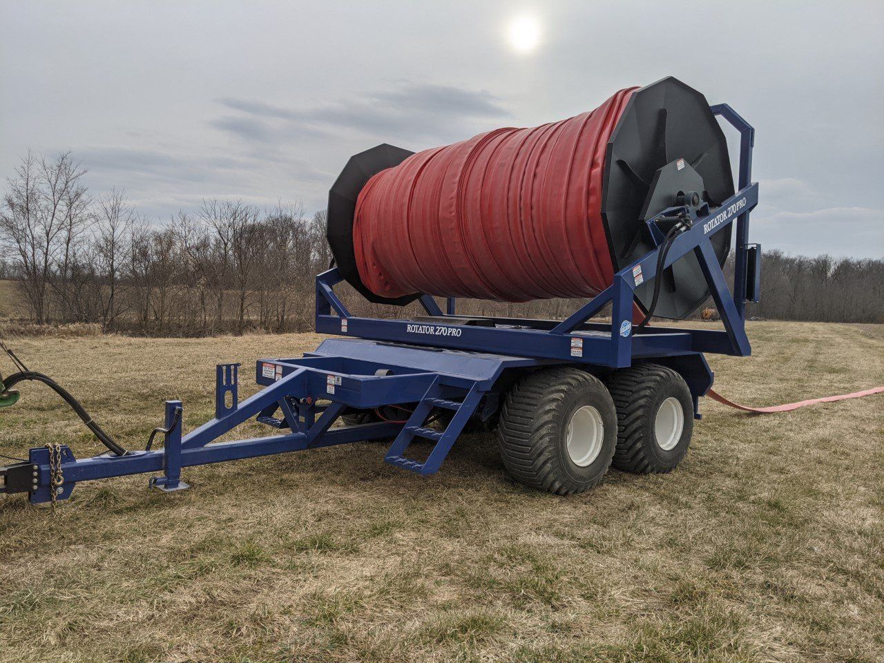 Blue farm trailer with a large red hose reel in a grassy field