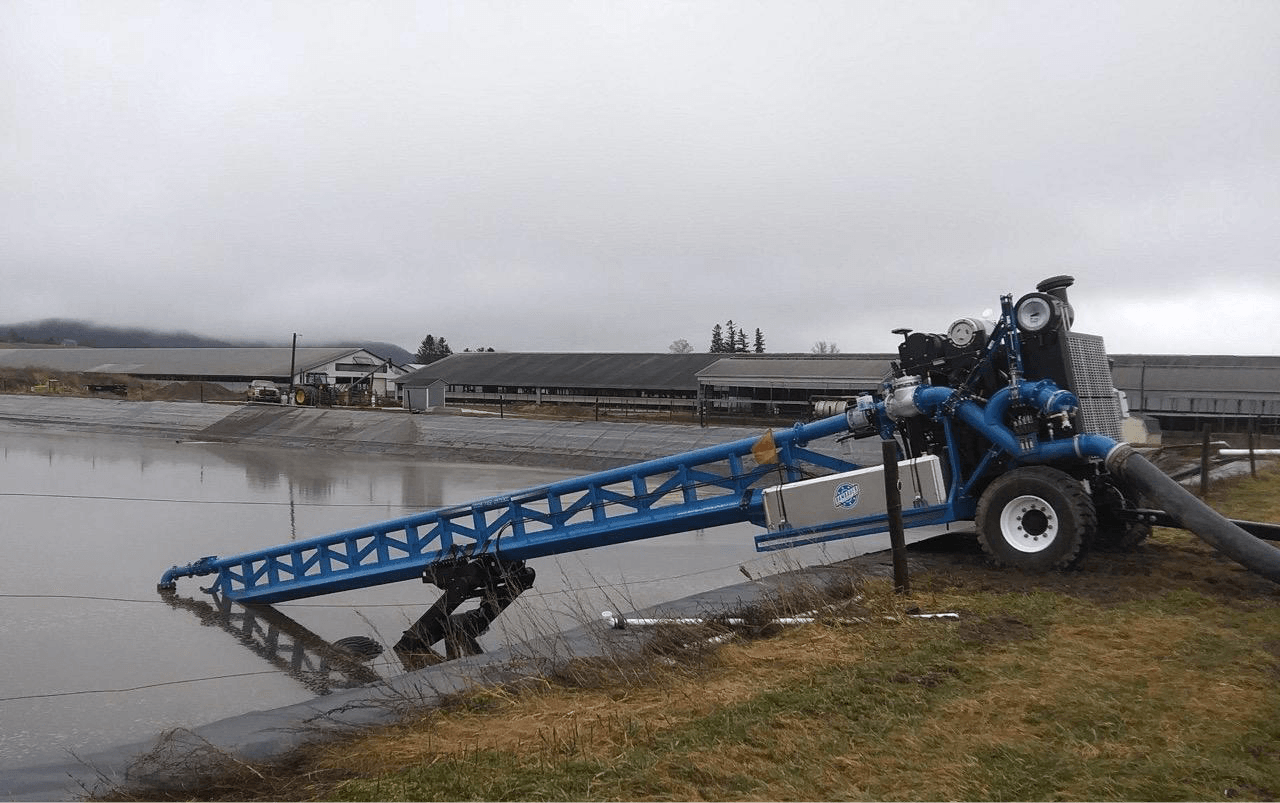 Blue agricultural machine draining water onto a muddy shoreline under an overcast sky