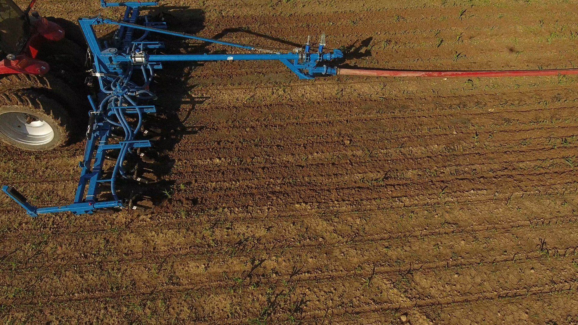 Blue farm machinery plowing a brown field, with a red pipe extending across the right side.