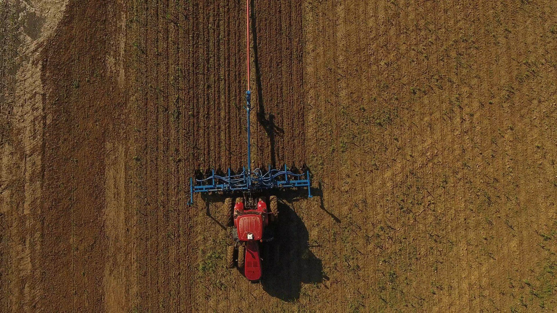 Red tractor plowing a brown field, viewed from above.