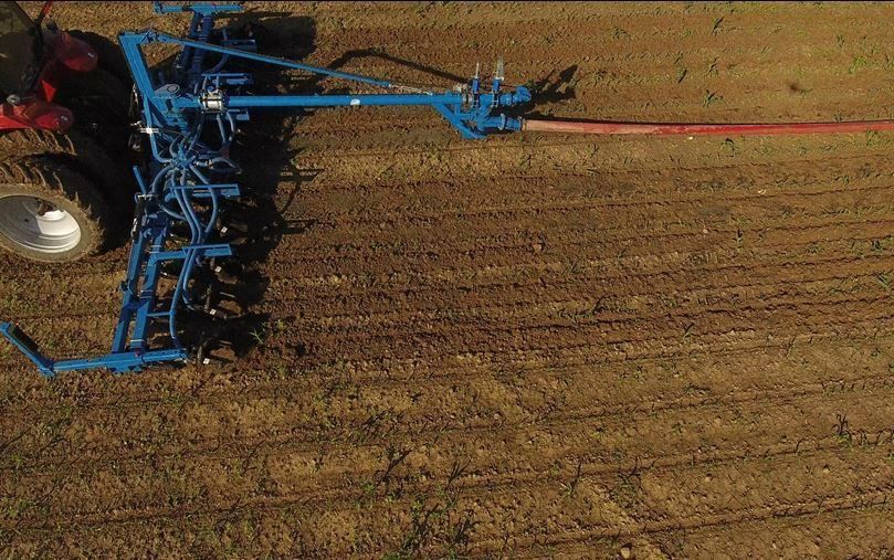 Blue farm tractor pulling a wide tillage implement across a brown field, viewed from above