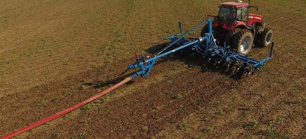 Red tractor pulling a blue agricultural implement across a brown field with a long hose attached