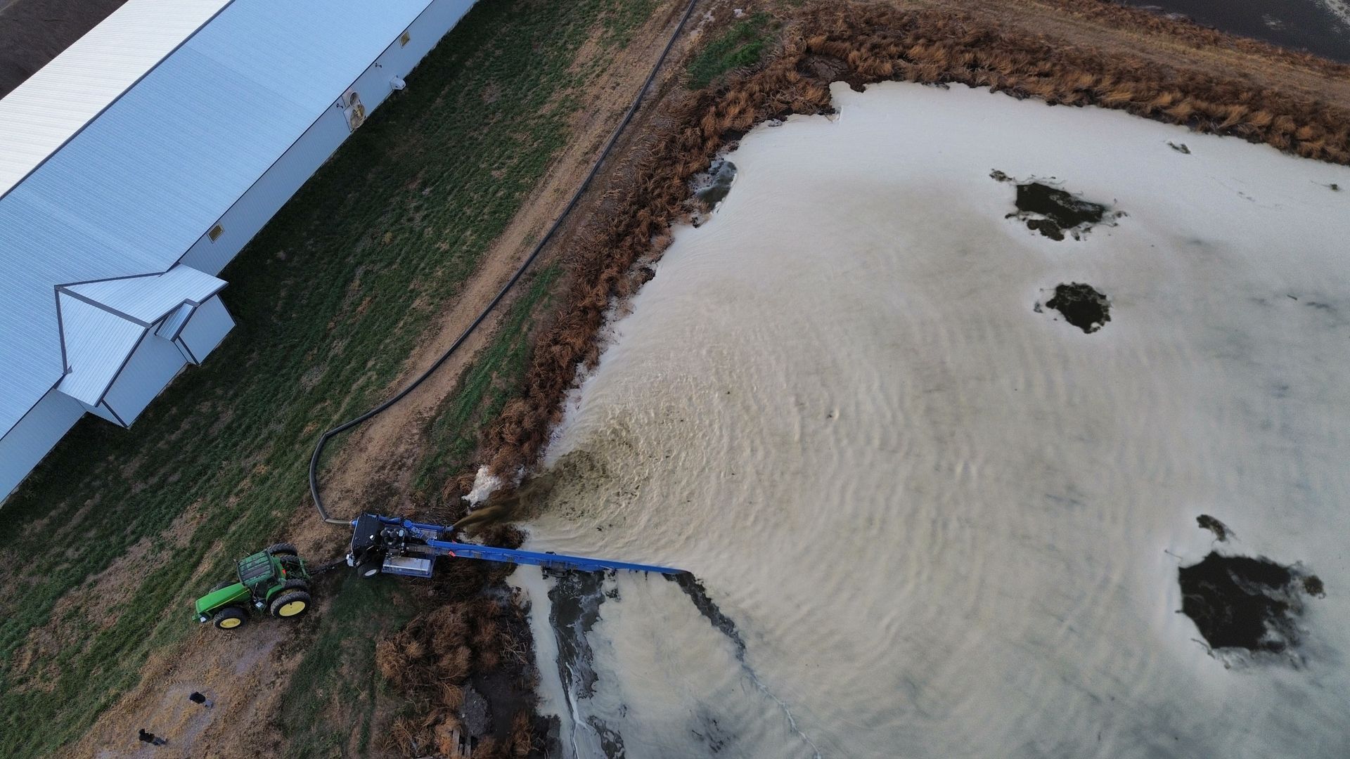 Aerial view of a green tractor spraying or pumping liquid into a pond beside a long building.