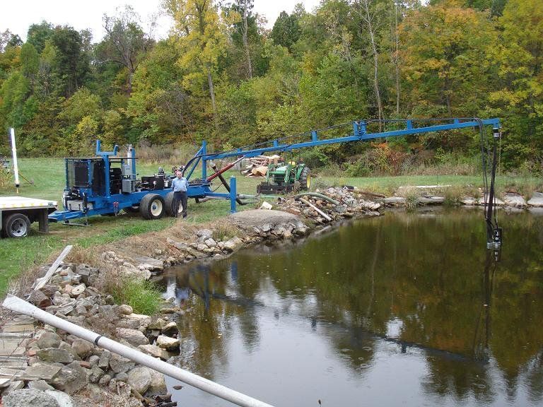 Blue pond-maintenance machine with long arm over a rocky shoreline and still water