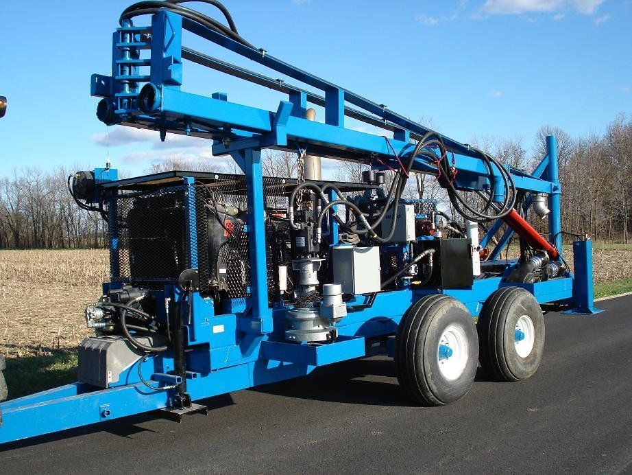Blue drilling rig trailer with large wheels and hydraulic arms in a field under a clear sky