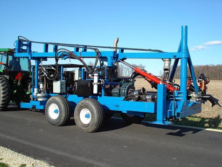Blue agricultural drilling rig on a trailer being towed along a road in a rural field.