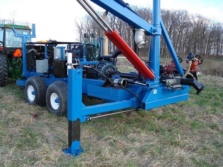 Blue agricultural machine on wheels in a grassy field, with a red hydraulic arm and two people nearby