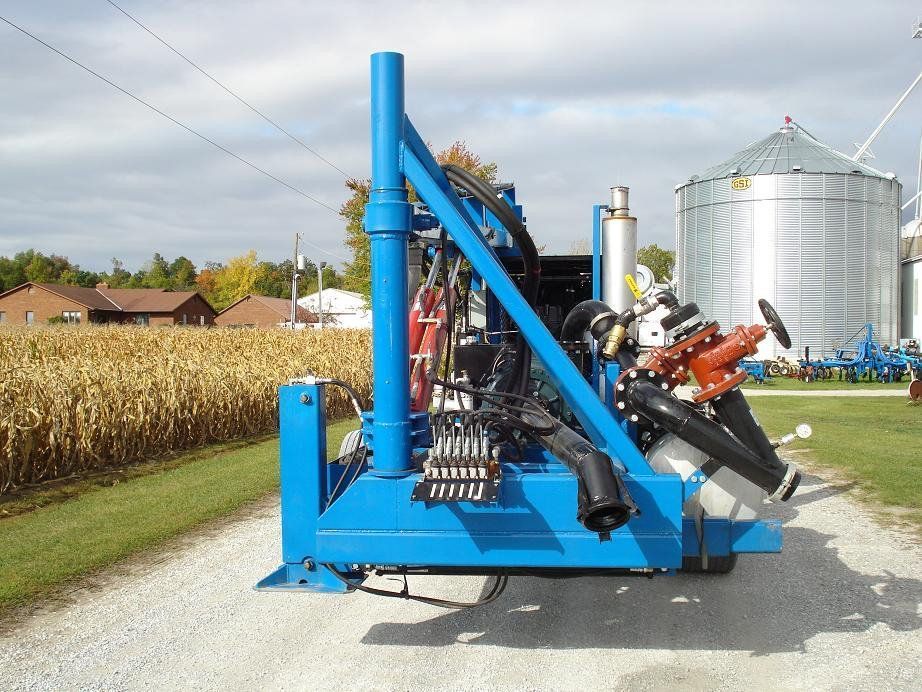 Blue farm implement parked beside a cornfield and silos on a gravel farm lane.