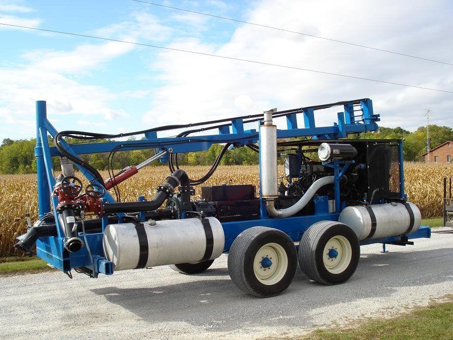 Blue agricultural sprayer trailer with tanks and hoses parked beside a cornfield