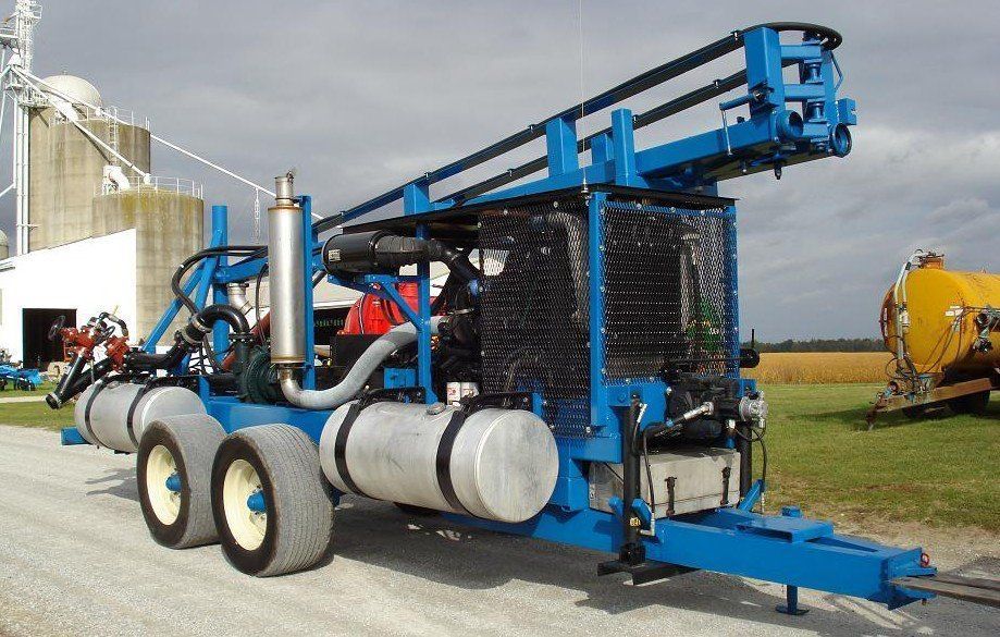 Blue farm machine trailer with large spools and hydraulic arm parked beside a building outdoors