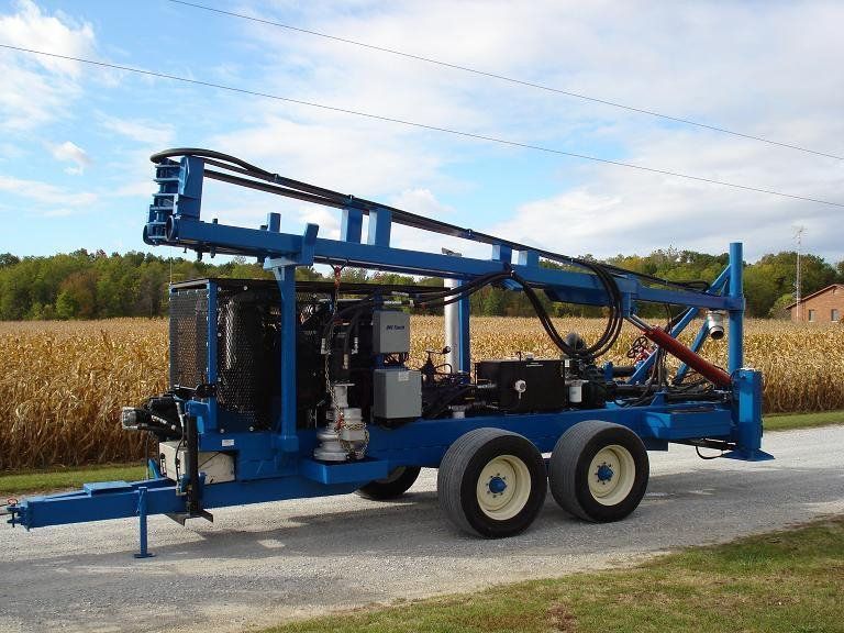 Blue drilling rig trailer on a rural road beside cornfields under a partly cloudy sky