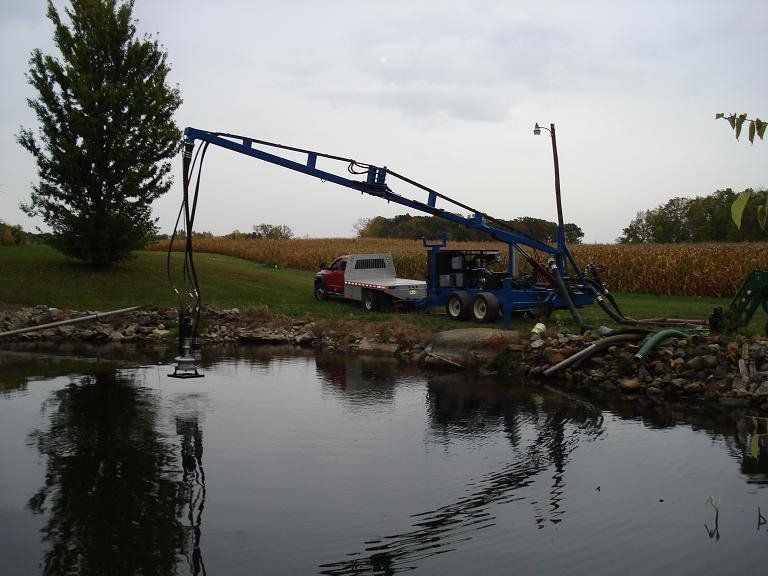 Blue drilling rig beside a pond with a white pickup truck and trees on a rural field edge