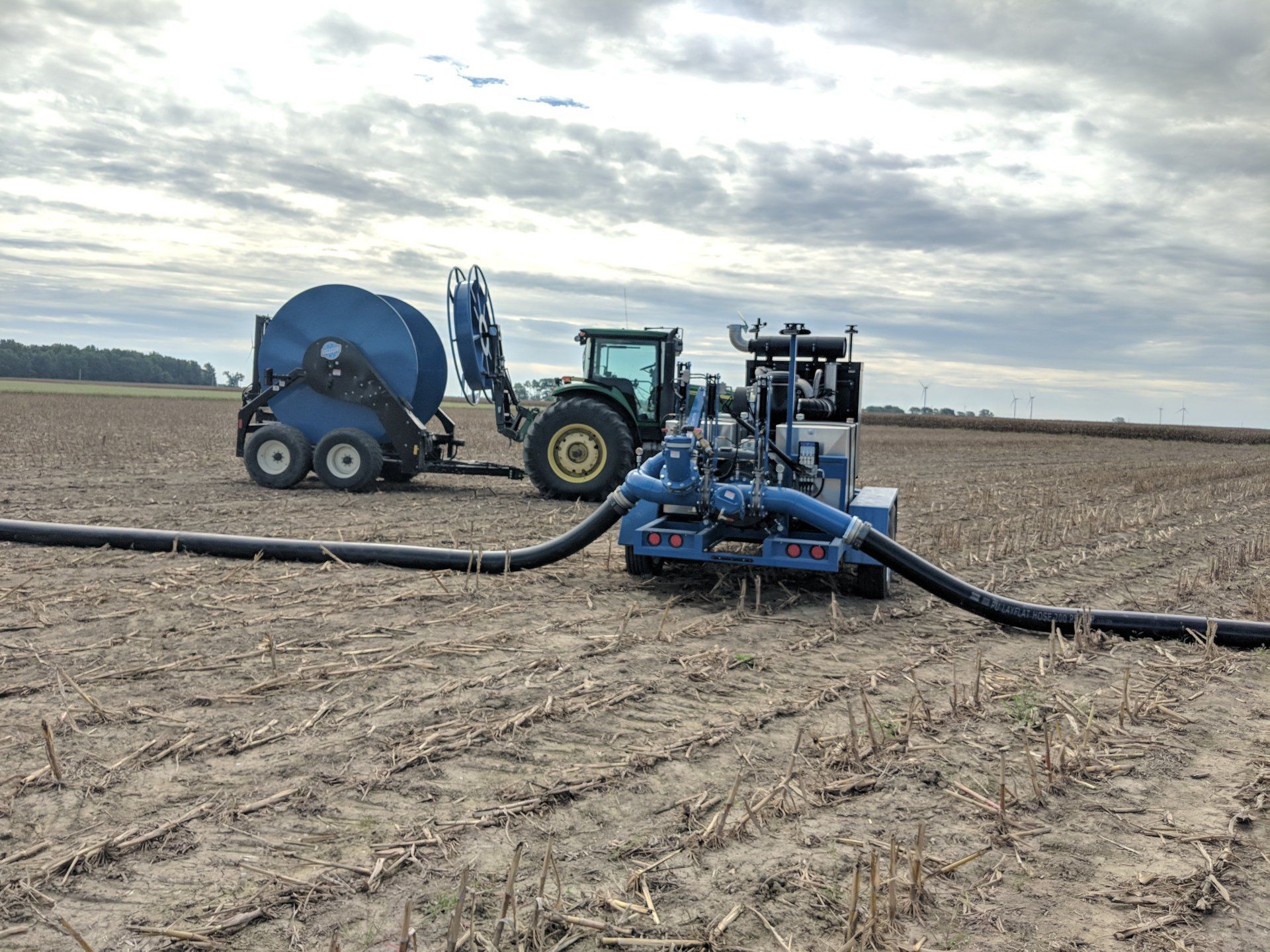 Farm machinery laying black irrigation pipe across a plowed field under a cloudy sky