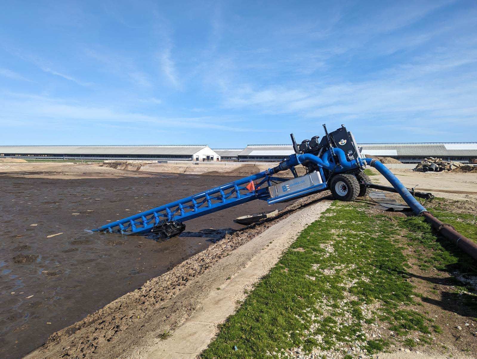 Blue farming machine tipped over beside muddy field and grassy embankment under a clear sky