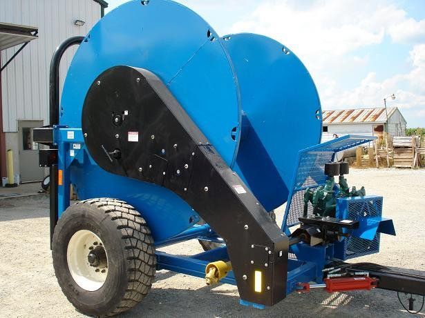 Blue agricultural machine on wheels with a black chute, parked outdoors near a building.