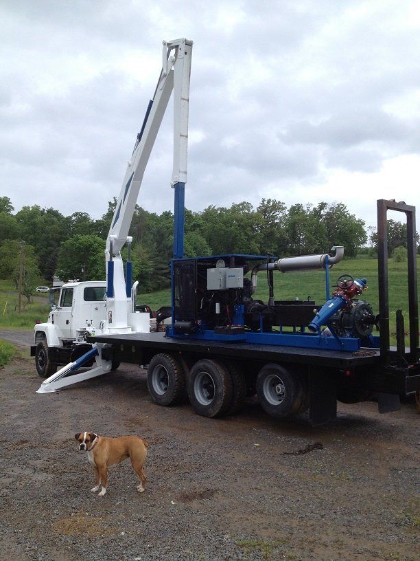 White utility truck with crane and equipment on a gravel lot; a brown dog stands in front.