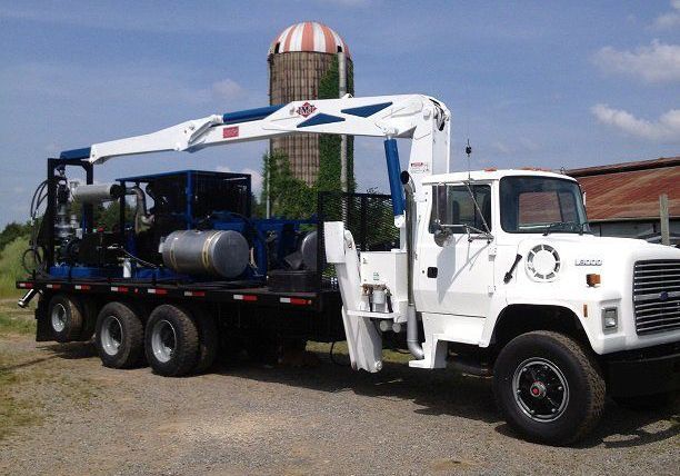 White flatbed truck with a crane carrying machinery in a gravel lot, with a silo in the background