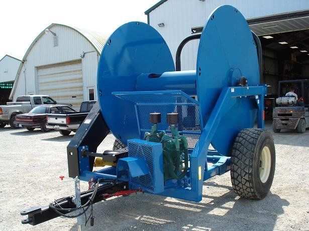 Blue industrial machine on a trailer in a farmyard, with large curved wheels and a warehouse in the background