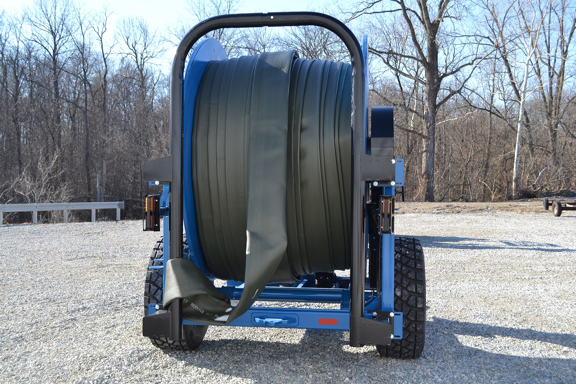Large blue hose reel trailer with coiled hose in a gravel yard, with bare trees in the background