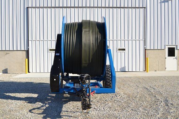 Blue hose reel trailer with large black hose, parked on gravel in front of a metal building.