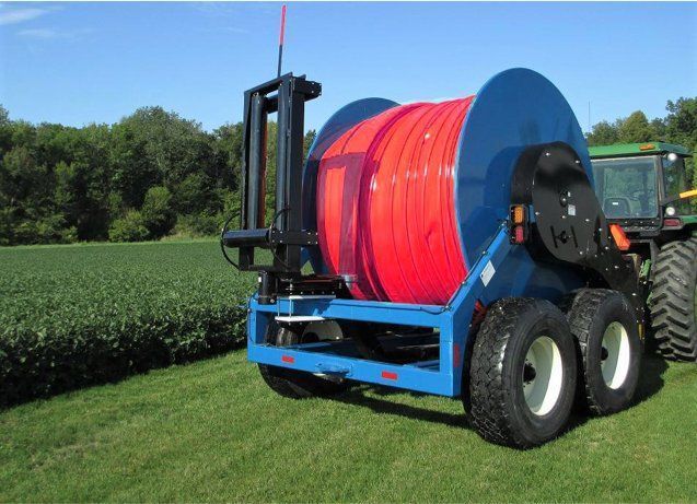 Blue farm irrigation reel sprayer with red hose parked beside crop rows in a green field