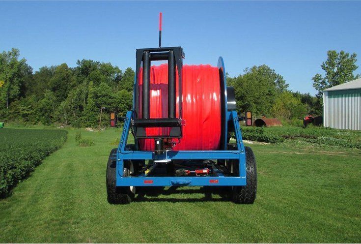 Blue hose reel trailer with bright red hose on a grassy field near trees and a barn