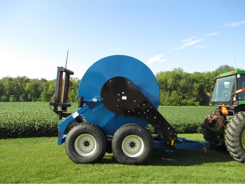 Blue agricultural reel trailer attached to a tractor in a field, with trees and sky in the background