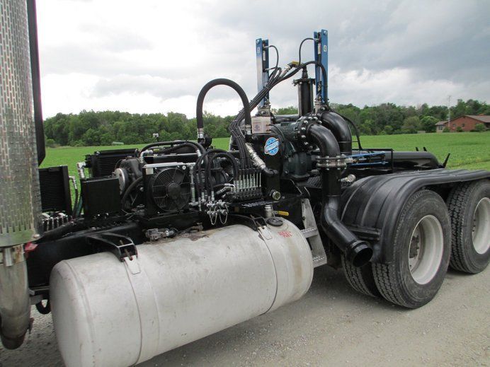 Black truck tractor with large white fuel tank parked in a grassy field under cloudy sky