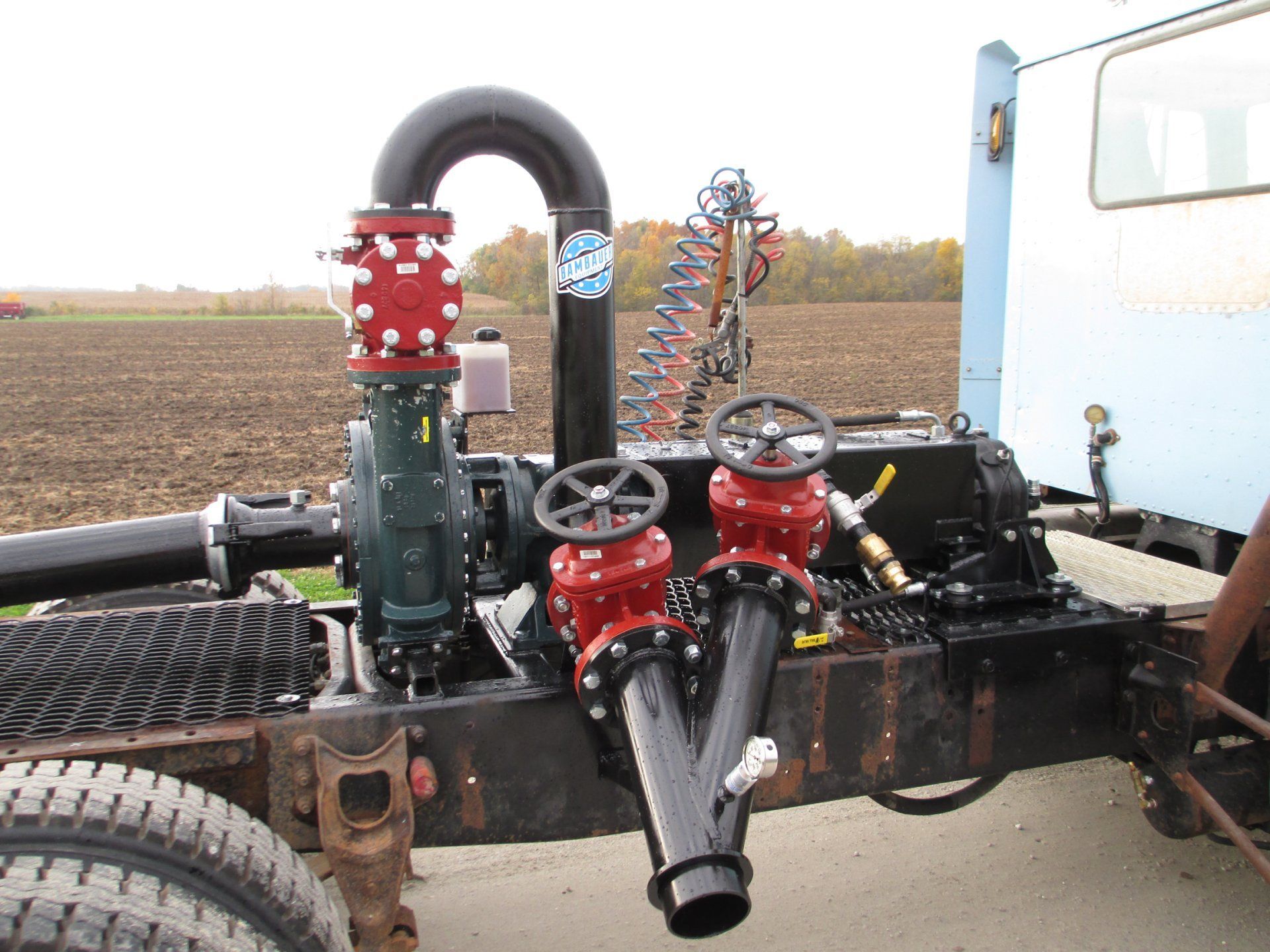 Industrial vehicle frame with red valves, black hoses, and a large tire in a field scene